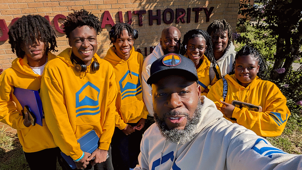 Group of young people and an adult posing for a selfie in front of the Palatka Housing Authority sign, all wearing yellow sweatshirts with a logo.